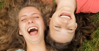 Two young women laughing on grass, eyes closed.